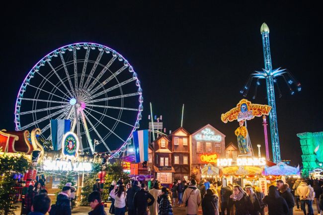 Illuminated big wheel, Santa Land sign and wooden chalets at Hyde Park Winter Wonderland at night, with crowds along the paths